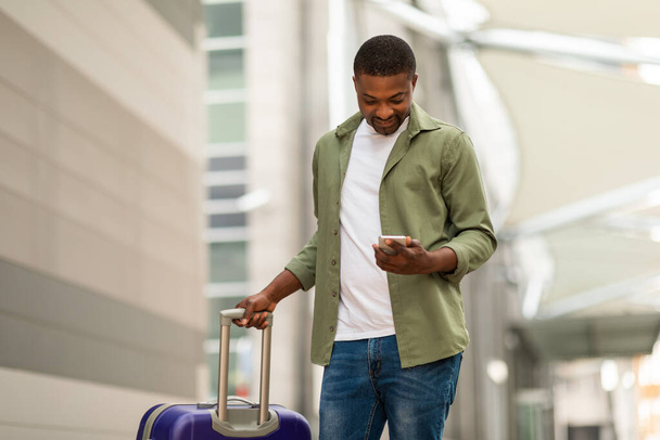 Счастливый чернокожий мужчина, пользующийся телефоном с туристическим приложением и стоящий с чемоданом в аэропорту. Парень бронирует билеты онлайн на мобильный телефон. Гаджеты и мобильные услуги для туризма - Фото, изображение