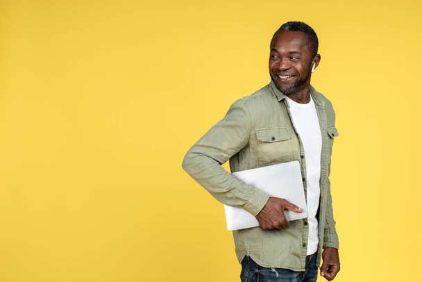 Smiling confident mature black male in casual, wireless headphones with laptop looking back at empty space, isolated on yellow background, studio. Great ad and offer, technology for work and business - Φωτογραφία, εικόνα