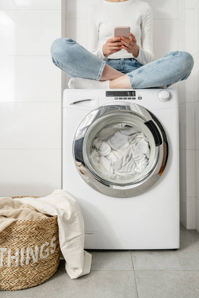 female housewife sitting with mobile phone on automatic washing machine loaded bedding and closed door. wicker basket with linen textile on floor in bathroom - Фото, зображення