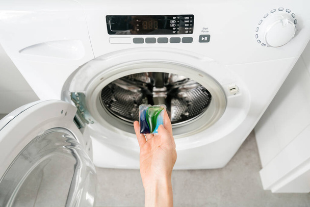 vrouwelijke hand houden wasmiddel capsule peulen in de buurt van automatische wasmachine met open deur in de badkamer, wasserette concept, bovenaanzicht - Foto, afbeelding