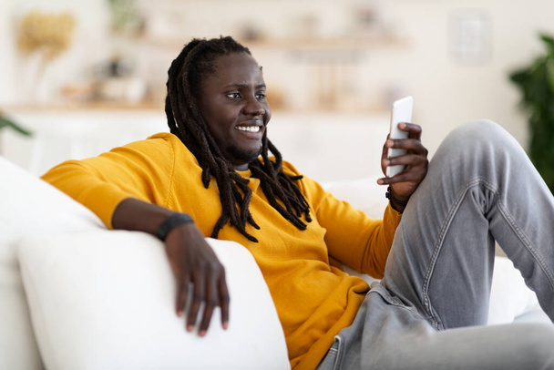 retrato de alegre preto millennial cara descansando com smartphone em casa, feliz jovem afro-americano homem relaxante no sofá na sala de estar, mensagens com amigos ou navegar na Internet, close-up - Foto, Imagem