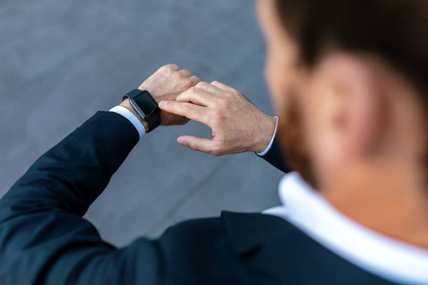 Young caucasian guy with beard in suit checks time on smartwatch with blank screen on street, unrecognizable. Businessman and business meeting, lifestyle, finance and startup, date and work control - Фото, изображение
