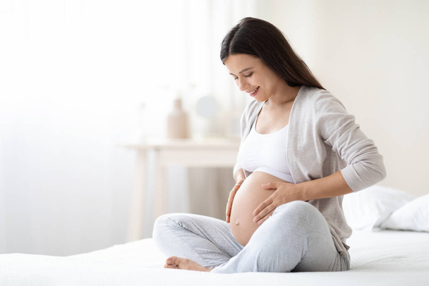 Smiling young pregnant woman sitting on bed in white bedroom at home, hugging her big tummy, feeling joyful as emotional well affect to baby neurological and psychological development, empty space - Photo, image