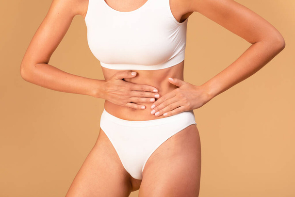 Young woman in white underwear touching left abdominal side, suffering from strong pain, beige studio background, cropped, closeup - Φωτογραφία, εικόνα