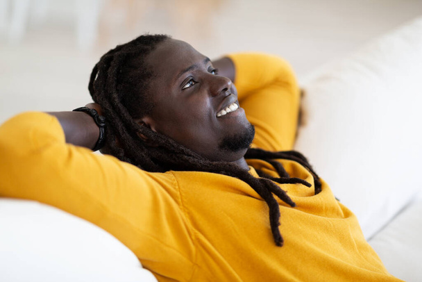 Ruhe zu Hause. Porträt eines lächelnden jungen Schwarzen, der sich auf der Couch zurücklehnt, verträumter Afroamerikaner, der auf dem Sofa sitzt, die Hände hinter dem Rücken, Millenial Guy, der sich im Wohnzimmer entspannt, Nahaufnahme - Foto, Bild