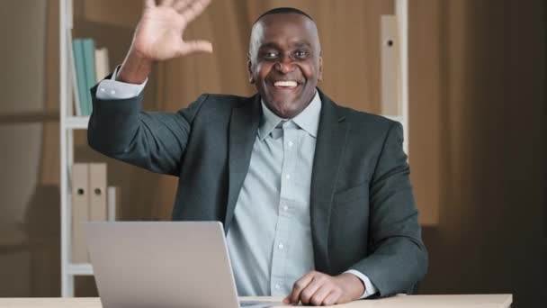 African American old senior businessman boss worker man sitting at table in office with laptop looking at camera waving hands hi hello greeting happy showing thumbs up talking video call conference - Footage, Video