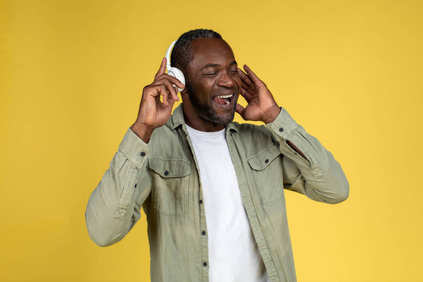 Tevreden volwassen zwarte man in casual met draadloze hoofdtelefoon met gesloten ogen genieten van muziek, zingen alleen, plezier hebben, geïsoleerd op gele achtergrond, studio. Entertainment met audio app en lifestyle - Foto, afbeelding