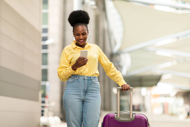 スーツケース付きのスマートフォンスタンドを使用してハッピーブラック観光女性空港ターミナル屋外でモバイルアプリケーションを介して旅行チケットを予約します。観光とガジェットのコンセプト - 写真・画像