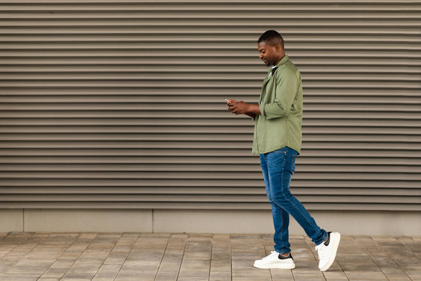 Cheerful African American Man Using Smartphone Texting Walking In Urban Area In City Outdoors. Millennial Guy Using New Mobile Application Outside. Communication And Gadgets Lifestyle Concept - Foto, imagen