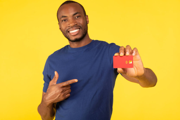 Toothy чорний чоловік показує червону кредитну картку для реклами великого банку Пропозиція вказуючи пальцем позує на жовтому фоні студії. Концепція фінансового та банківського обслуговування. Вибірковий фокус
 - Фото, зображення