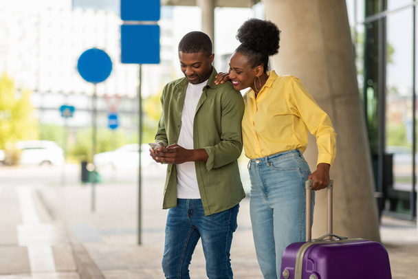 モバイルアプリケーションで携帯電話予約便を利用しているハッピーブラック観光客は、空港ターミナルの屋外近くに旅行スーツケースを持って立ちます。観光とモバイルコミュニケーションのコンセプト - 写真・画像