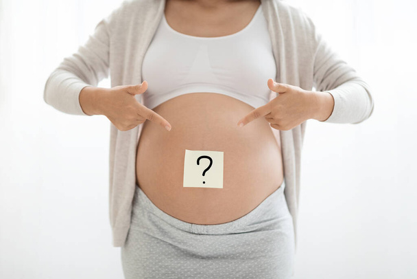 Ausschnitt einer werdenden Dame in Homewear mit Papieraufkleber mit Fragezeichen auf ihrem großen Bauch, weißer Hintergrund, unkenntliche Schwangere, die das Geschlecht des Babys erraten hat - Foto, Bild