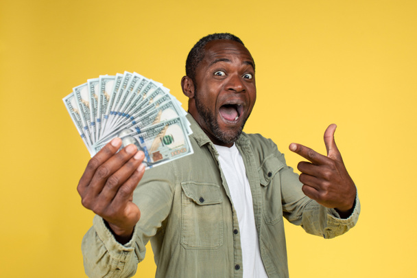 Счастливый шокирован смешной взрослый африканский американец мужчина в случайном с открытым ртом указывая пальцем на многие доллары в руке, изолированные на желтом фоне, студия. Выражение лица, победа, наличные и финансы - Фото, изображение