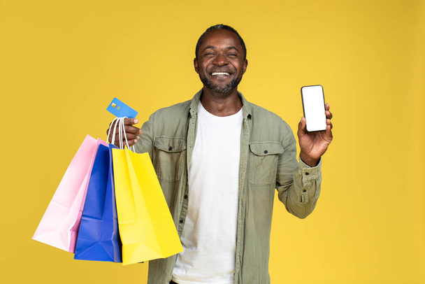 Счастливый взрослый африканский американец в случайном показе смартфона с пустым экраном и много сумок с покупками изолированы на желтом фоне, студия. Приложение для онлайн-покупок, продаж, банковского дела и финансов - Фото, изображение