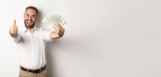 Excited man showing thumbs up and money, earning cash, standing over white background. - Photo, Image