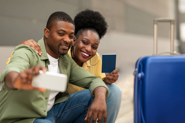 休暇旅行。空港でスーツケースとパスポートで携帯電話のポーズでセルフィーを作る幸せなアフリカ系アメリカ人観光客カップル。観光とモバイル技術。選択的フォーカス - 写真・画像
