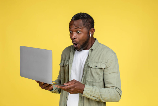 Surpreendido feliz adulto engraçado afro-americano masculino em casual e fones de ouvido olhando para laptop, isolado em fundo amarelo, estúdio. As pessoas emoções, sucesso e vitória, anúncio chocante, venda e oferta - Foto, Imagem