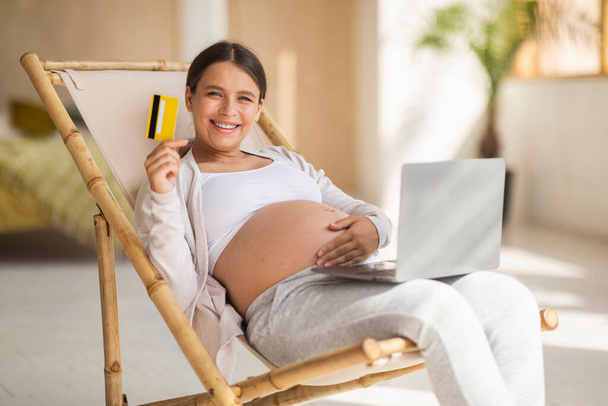İnternetten alışveriş. Dizüstü bilgisayarı ve kredi kartı olan mutlu ve hamile bir kadın koltukta dinleniyor, gülümseyen bir kadın evde bilgisayarla dinlenirken internet alışverişi yapıyor, - Fotoğraf, Görsel