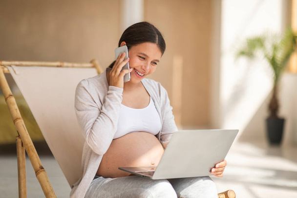 Wolny strzelec w czasie ciąży. Młoda kobieta w ciąży pracująca z laptopem i rozmawiająca na komórce siedząc na krześle w domu, zadowolona z pracy zdalnej, przestrzeń kopiująca - Zdjęcie, obraz