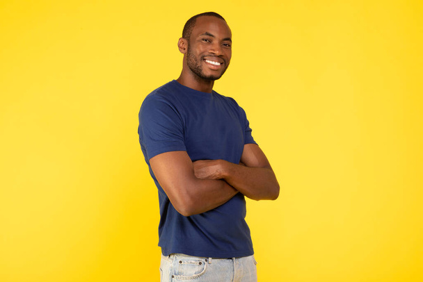 Cheerful African American Man Posing Smiling To Camera Standing Crossing Hands Over Yellow Studio Background. Shot Of Cheerful Black Guy. Male Beauty Concept - 写真・画像