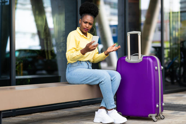 Niezadowolony African American Woman Tourist Using Smartphone having problem Rezerwacja biletów online siedzi z walizką w obszarze miejskim na zewnątrz. Koncepcja zagadnień związanych z podróżami i komunikacją - Zdjęcie, obraz