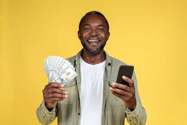 Satisfied adult african american male in casual hold many dollars and smartphone, isolated on yellow background, studio, free space. Emotions from online win, app for cashback, finance and savings - Фото, зображення