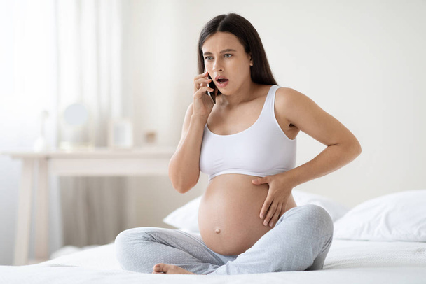 Shocked young pregnant woman in homewear sitting on bed, touching her big tummy and having phone conversation, feeling some pain and discomfort at home, calling doctor, panorama with copy space - Photo, image