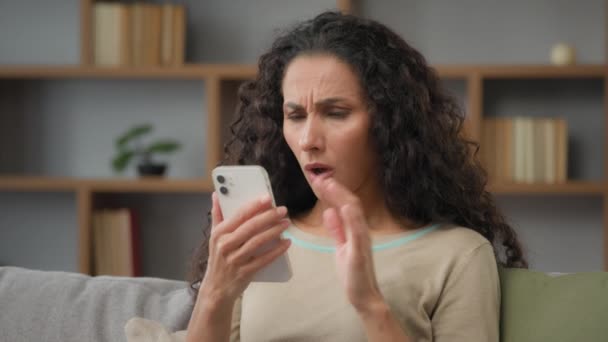 Hispanic sad displeased woman has problem with online mobile app on cell phone unhappy with blocked account in social networks looking at cellphone screen scam unsuccessful bad news sitting at home - Filmmaterial, Video