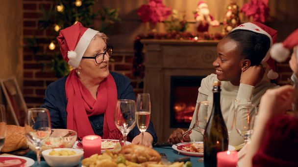 Senior woman talking with festive young adult person while enjoying Christmas dinner at home. Multicultural people drinking champagne while celebrating winter hoilday together. - Foto, Imagem