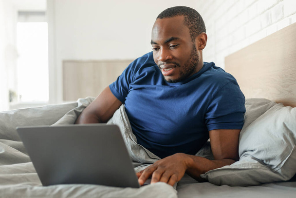 Black Freelancer Guy Using Laptop Computer Working Online Lying In Modern Bedroom At Home. Man Browsing Internet In Bed On Weekend. Technology And Rermote Work Concept - 写真・画像