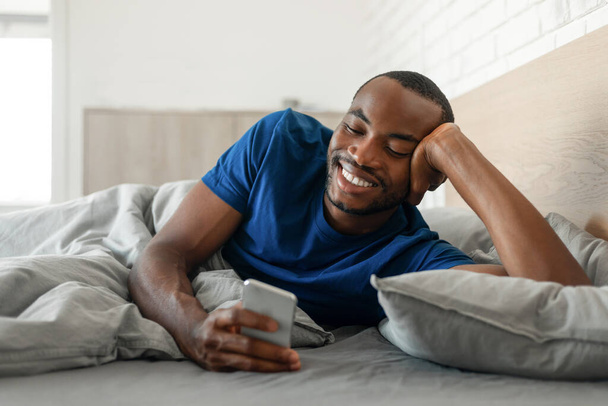 Ευτυχισμένος μαύρος τύπος που χρησιμοποιεί το κινητό τηλέφωνο στο κρεβάτι γραπτών μηνυμάτων και Websurfing το πρωί ανάπαυσης στη σύγχρονη κρεβατοκάμαρα στο σπίτι. Gadgets και έννοια επικοινωνίας. Επιλογή εστίασης - Φωτογραφία, εικόνα