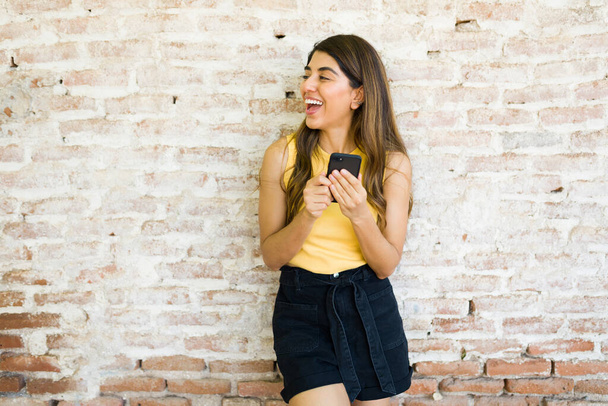 Iloinen latino nainen nauraa tiiliseinän edessä ja pitää hauskaa älypuhelinta käytettäessä  - Valokuva, kuva