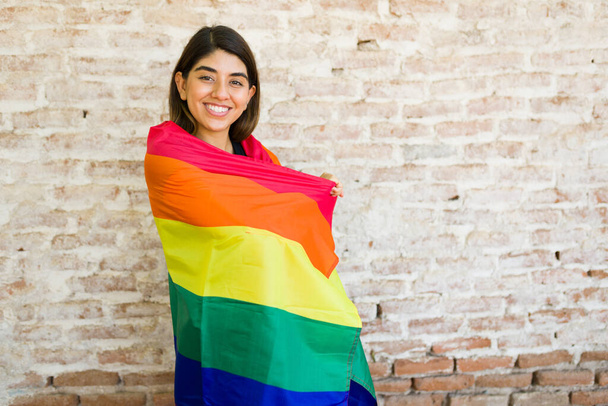 gai gay femme enveloppé dans un arc-en-ciel fierté drapeau et sentiment heureux tout en s'amusant devant un mur de briques à l'extérieur  - Photo, image