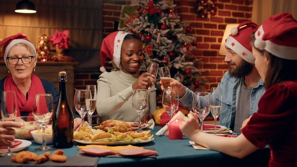 クリスマスディナーで自信のある男性とシャンパングラスをクリンクするアフリカ系アメリカ人女性。冬の休日を一緒に祝いながら伝統的な家庭料理を楽しむ楽しい家族. - 写真・画像