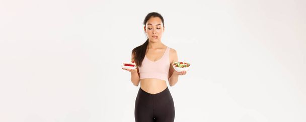 Active lifestyle, fitness and wellbeing concept. Portrait of indecisive and tempting cute asian girl trying resist temptation as looking at delicious cake, being on diet, need eat healthy salad. - Photo, Image