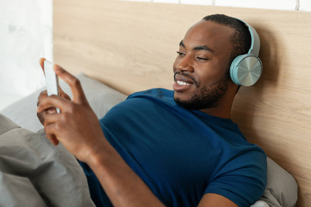 Relajado hombre negro escuchando música a través del teléfono con auriculares inalámbricos acostado en el dormitorio en casa por la mañana. Guy usando aplicación musical. Concepto de Tecnología y Gadgets - Foto, Imagen