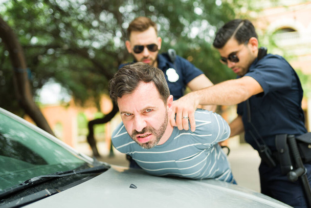 Wściekły młody człowiek wygląda na zdenerwowanego i agresywnego podczas aresztowania przez dwóch gliniarzy zakładających kajdanki za popełnienie przestępstwa - Zdjęcie, obraz