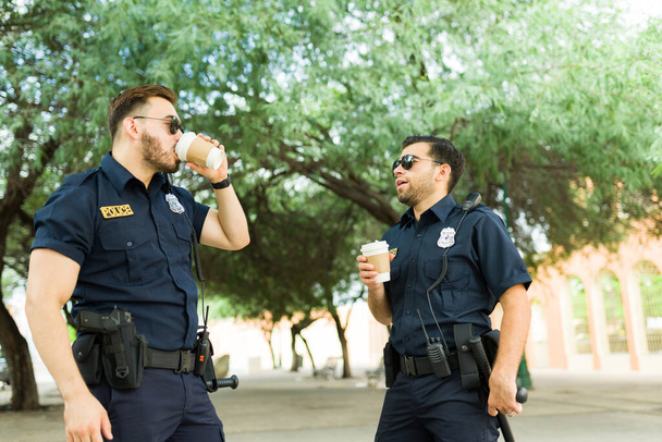 Relaxed police officers drinking coffee together and talking while on duty in law enforcement  - 写真・画像