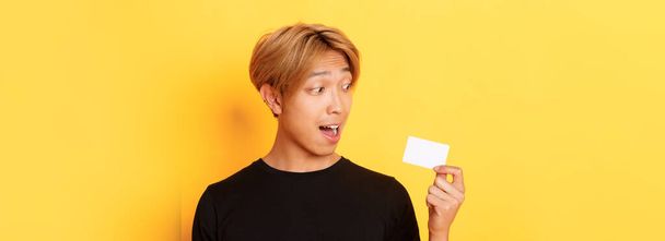 Close-up zdumiony Azji facet mówi o karcie kredytowej, stojąc nad żółtym tle. - Zdjęcie, obraz