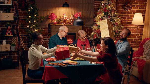 Joyful people gathered at Christmas dinner table while exchanging gifts. Multiethnic family members celebrating traditional winter holiday while giving each other presents. - Foto, Imagen