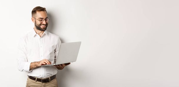 Zaken. Knappe zakenman die aan laptop werkt, berichten beantwoordt en lacht, over een witte achtergrond staat. - Foto, afbeelding