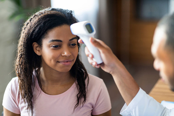 Крупним планом чорний чоловік лікар перевіряє пацієнта на досить афроамериканську температуру тіла, використовуючи електронний термометр, молода леді перед вакцинацією проти коронавірусу
 - Фото, зображення