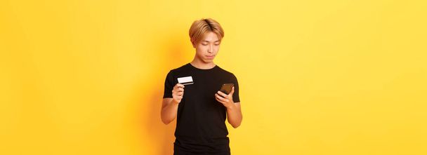 Young handsome asian guy in black t-shirt shopping online, holding credit card and using smartphone, standing yellow background. - Фото, изображение