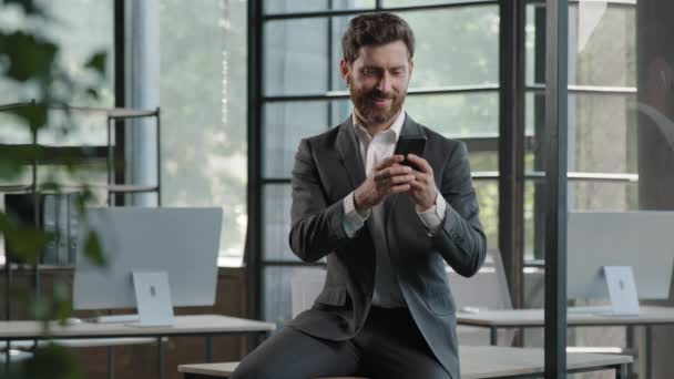携帯電話のチャットを使用してスマートフォンの画面を見てオフィスでテーブルの上に座って大人の会社の最高経営責任者(CEO)の労働者の上司のマネージャーの男の専門家のビジネスマンオンラインチェックデータ主催者 - 映像、動画