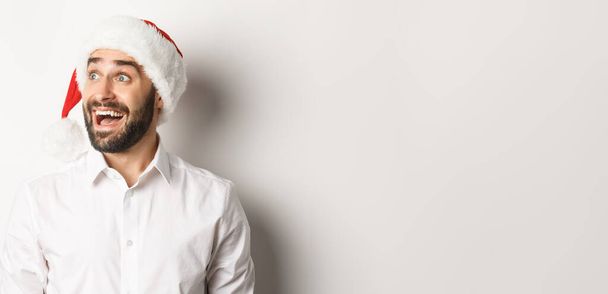Közelkép a csodálkozó szakállas férfi télapó kalap, nézte balról izgalom, karácsonyi promóciós ajánlat, áll a fehér háttér. - Fotó, kép