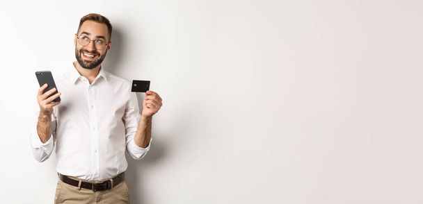 Business and online payment. Image of handsome man thinking while holding credit card and smartphone, standing against white background. - Foto, Imagem