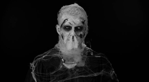 Frightening man with Halloween zombie bloody wounded makeup, trying to scare, blows smoke from nose and mouth. Horror theme. Sinister undead guy isolated on studio black background. Voodoo rituals - Photo, Image