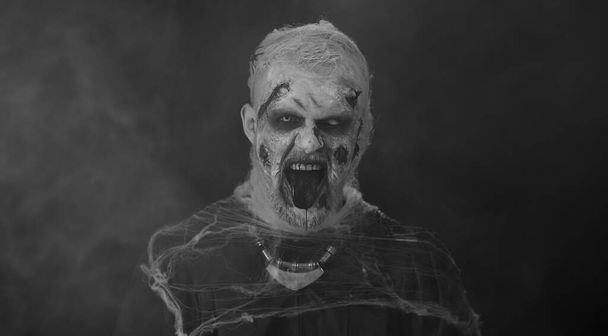 Złowrogi straszny człowiek w karnawałowym kostiumie szalonego zombie z krwawymi rannymi bliznami na twarzy, próbujący wystraszyć odizolowany na czarnym pokoju. Temat horroru cosplay rannych nieumarłych, bestii, potwora - Zdjęcie, obraz