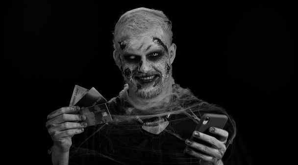 Sinister man with horrible scary Halloween zombie make-up using credit bank cards and smartphone while transferring money purchases online shopping. Dead guy with wounded bloody scars face, black room - Photo, Image