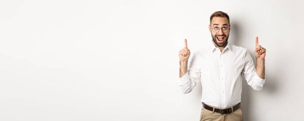 Excited businessman checking out advertisement, pointing and looking up with happy face, standing against white background. - Photo, Image